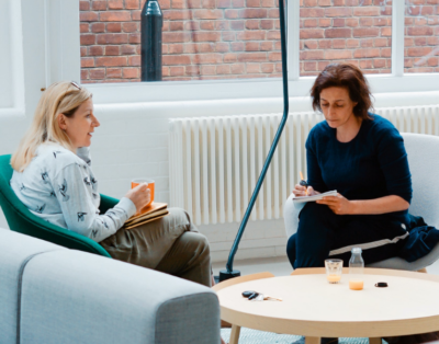 Untitled design (1) Two women sat facing each other talking, in front of them is a light brown coffee table.