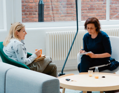 Two women sat facing each other talking, in front of them is a light brown coffee table.